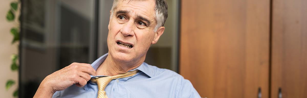 Mann in blauem Hemd und gelber Krawatte zieht gequält an seinem Kragen – Schweiß und Unwohlsein sind mögliche Symptome einer Unterzuckerung.
