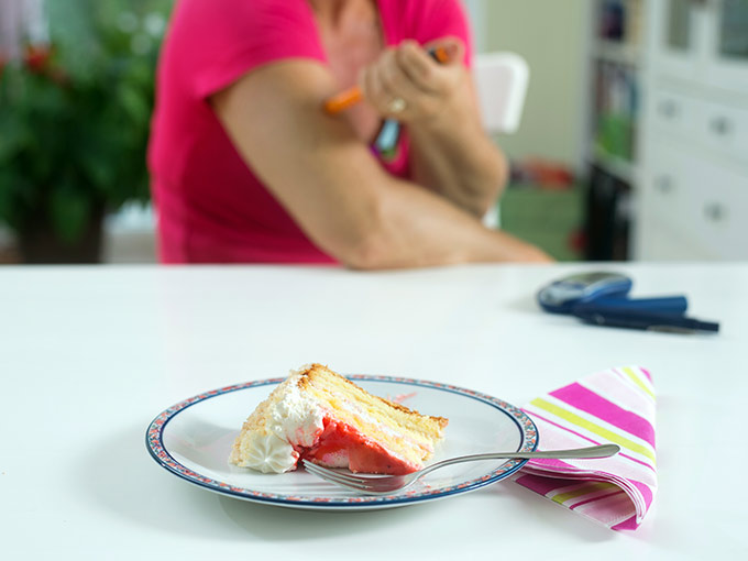 Im Vordergrund steht ein Stück Kuchen auf einem Tisch, im Hintergrund spritzt sich eine Diabetikerin Insulin - mit Diabetes ist es wichtig, keinen Korrekturfaktor zu kennen.