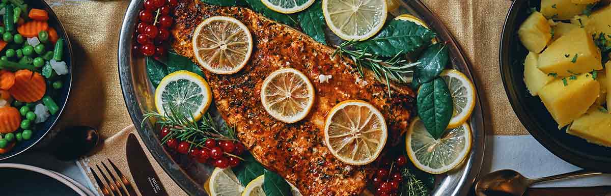 Fisch mit Zitronenscheiben garniert und neben Gemüsebeilagen auf einem Tablett - Fisch zählt zu den blutdrucksenkenden Lebensmitteln.